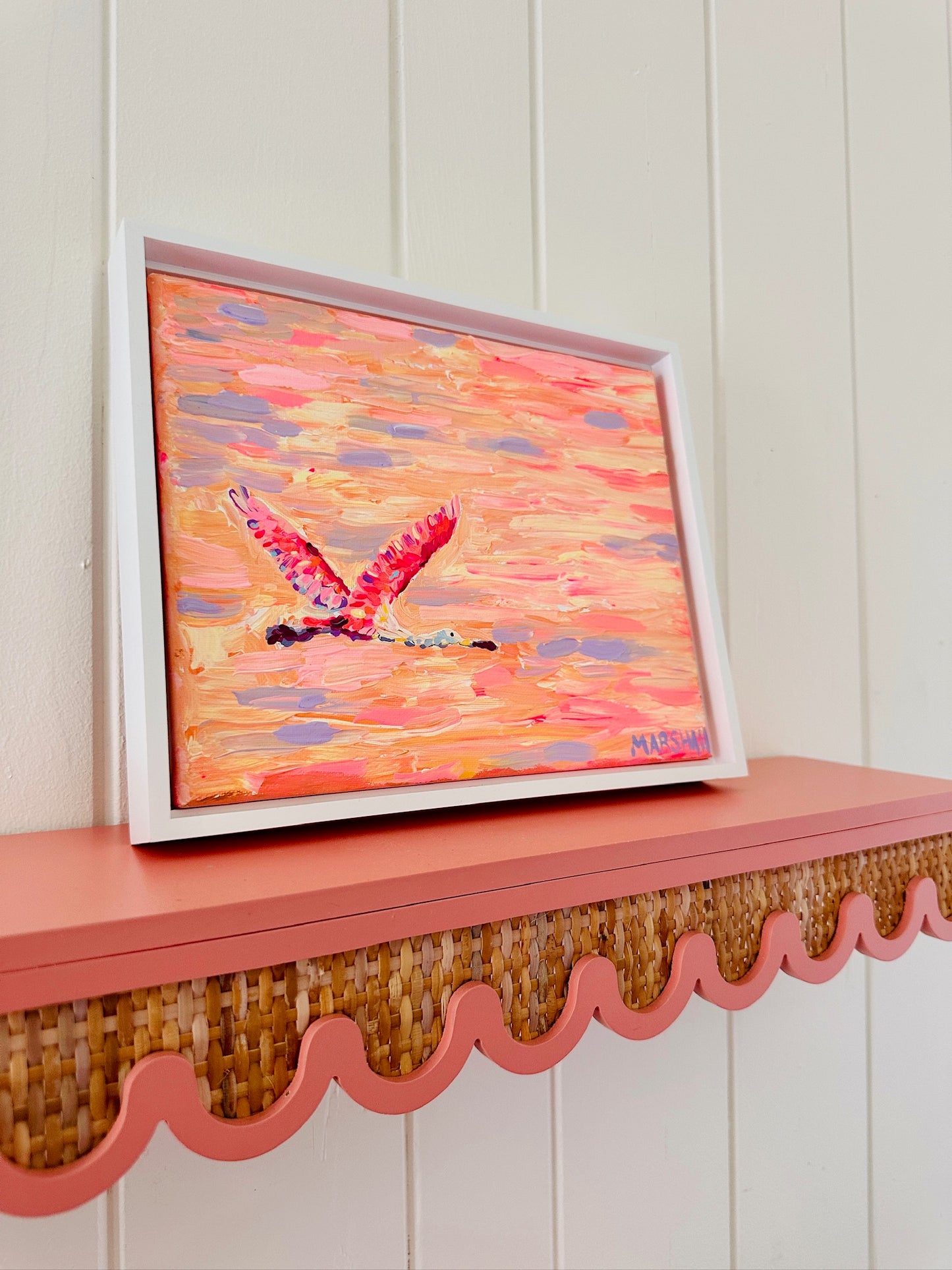 A Lauren Marshall painting of a colorful spoonbill in flight on a pink wavy shelf, set against a white paneled wall.