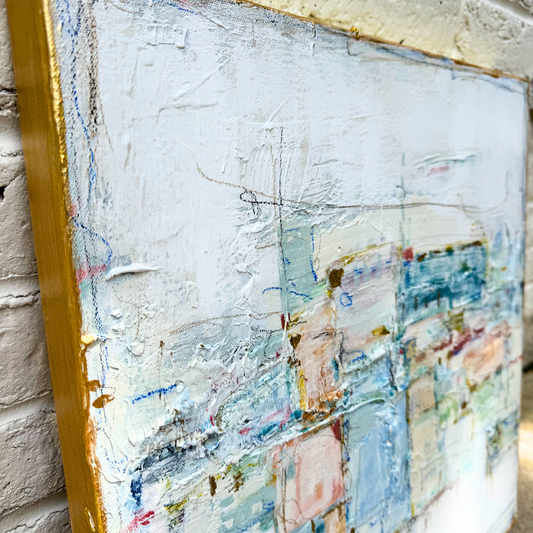 Close-up of a Megan Guidry abstract painting in blue pastels, set in a gold frame against a brick wall.