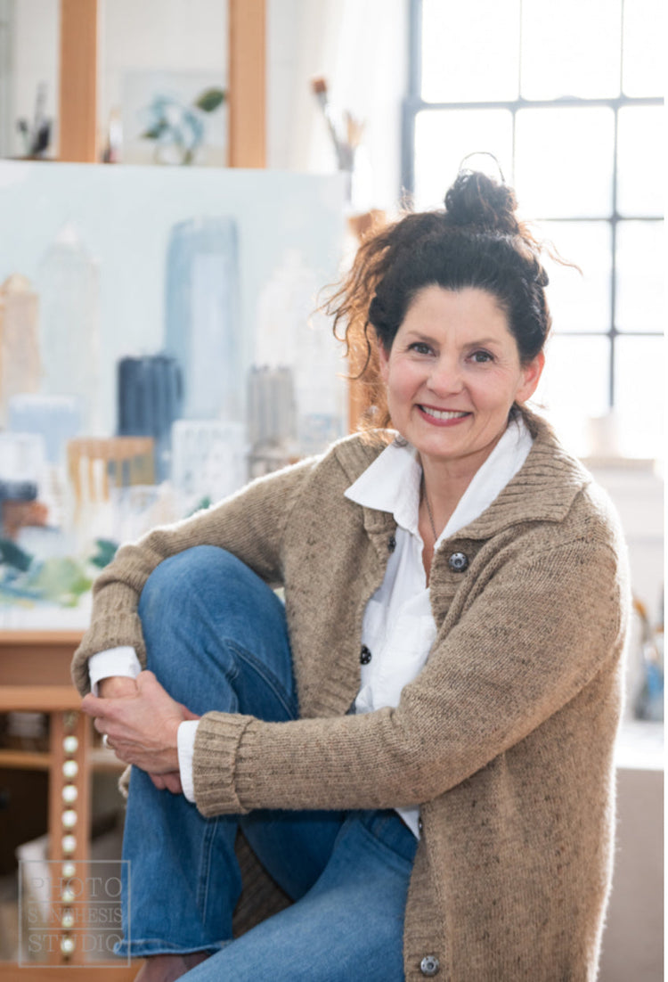 A woman with dark curly hair, wearing a beige sweater and jeans, sits smiling in an art studio with a cityscape painting and bright window in the background.