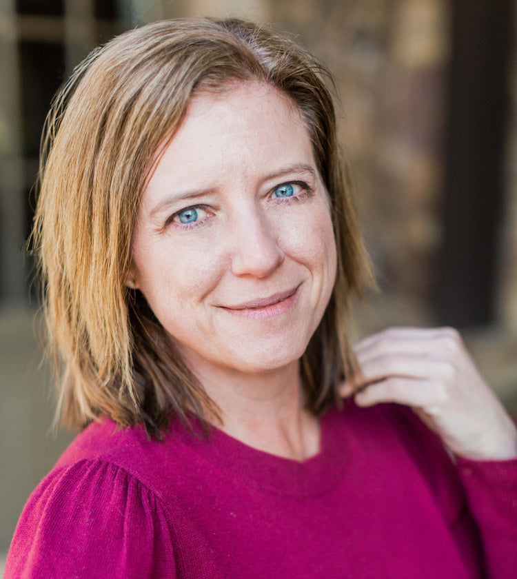 A woman with shoulder-length light brown hair and blue eyes smiles slightly, wearing a magenta top, standing in front of a blurred outdoor background.
