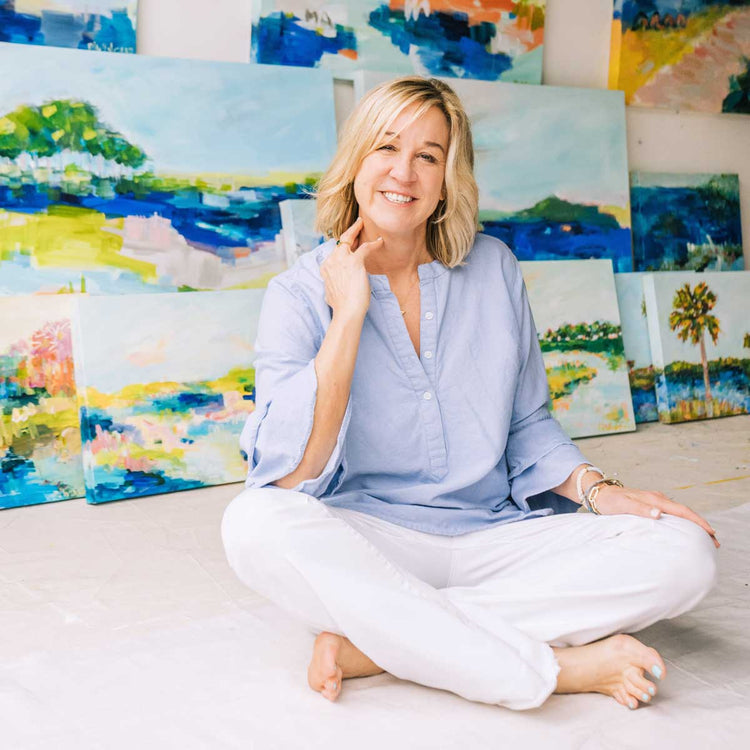 A woman in a light blue shirt and white pants sits barefoot on the floor, smiling, surrounded by colorful landscape paintings propped up behind her.