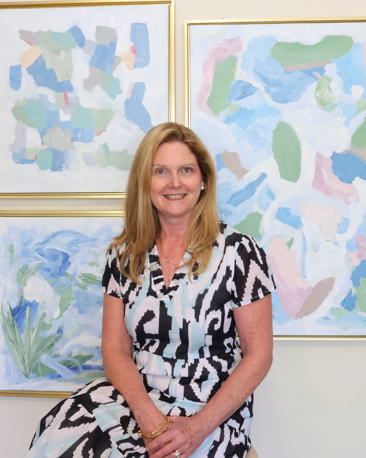 A woman with long blonde hair in a patterned dress sits smiling in front of three abstract pastel paintings in gold frames.