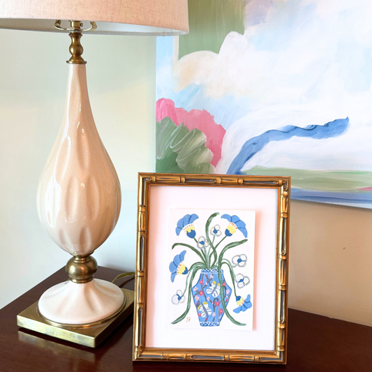 A table lamp and a framed botanical artwork sit on a wooden table, backed by a colorful original acrylic painting.