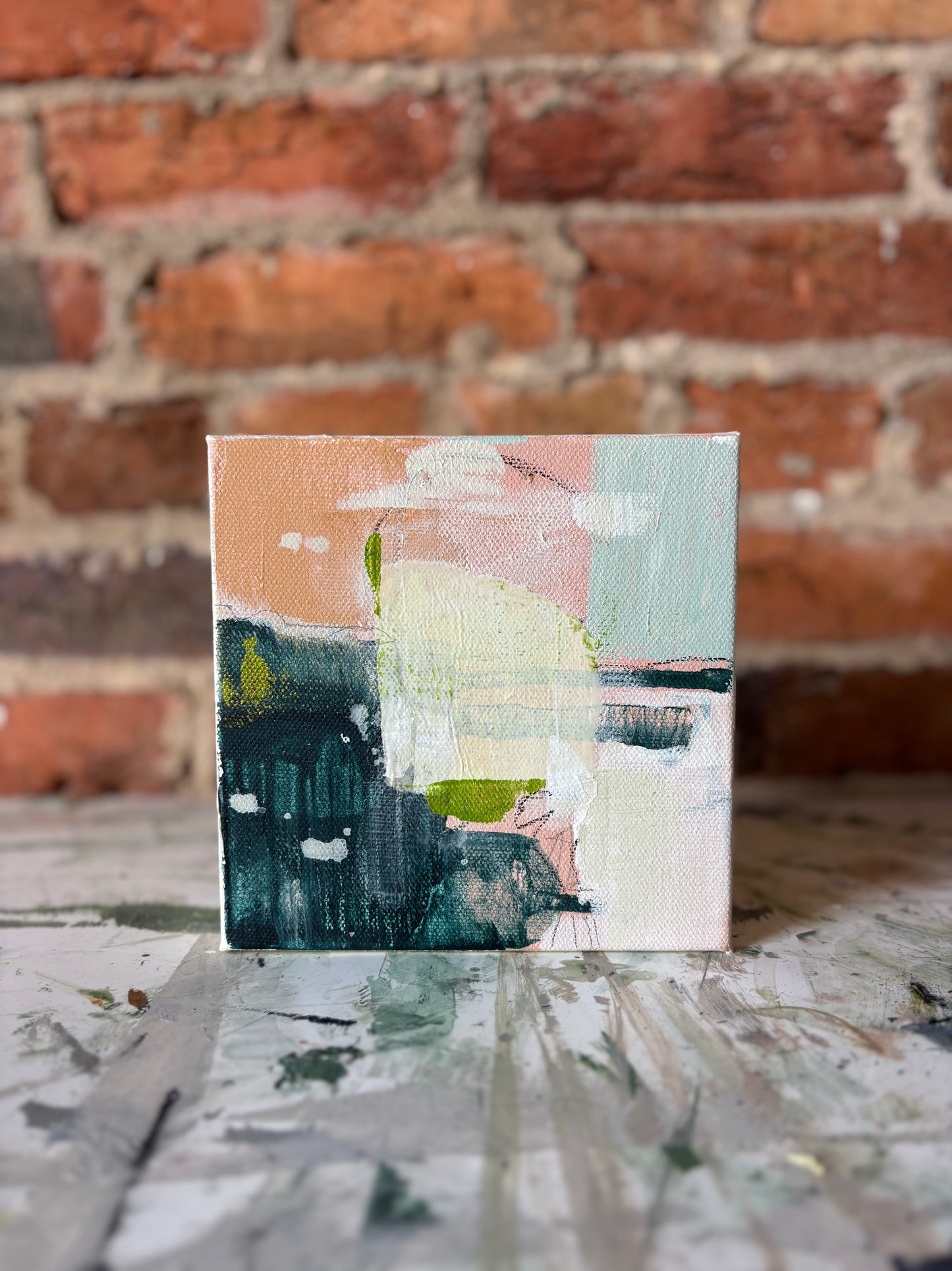 A small Emily Pullin mixed media artwork in pastel and dark tones rests on a paint-splattered surface by a brick wall.