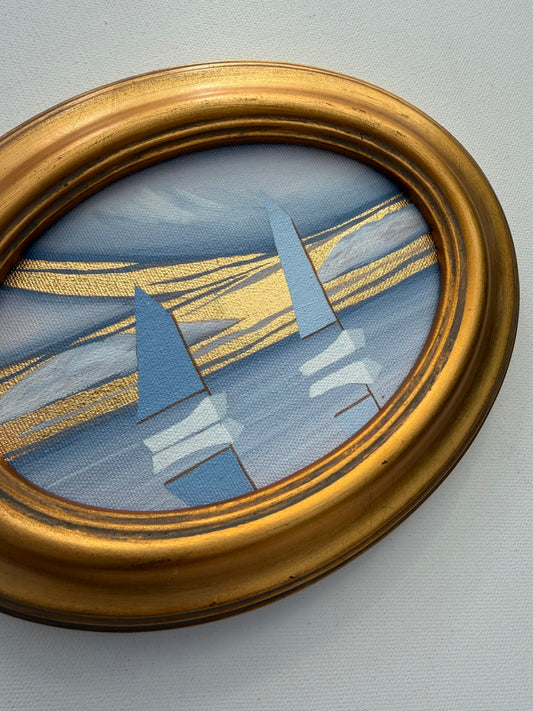 Oval coastal artwork in a gold frame depicting two blue sailboats on a light blue sea with gold streaks.