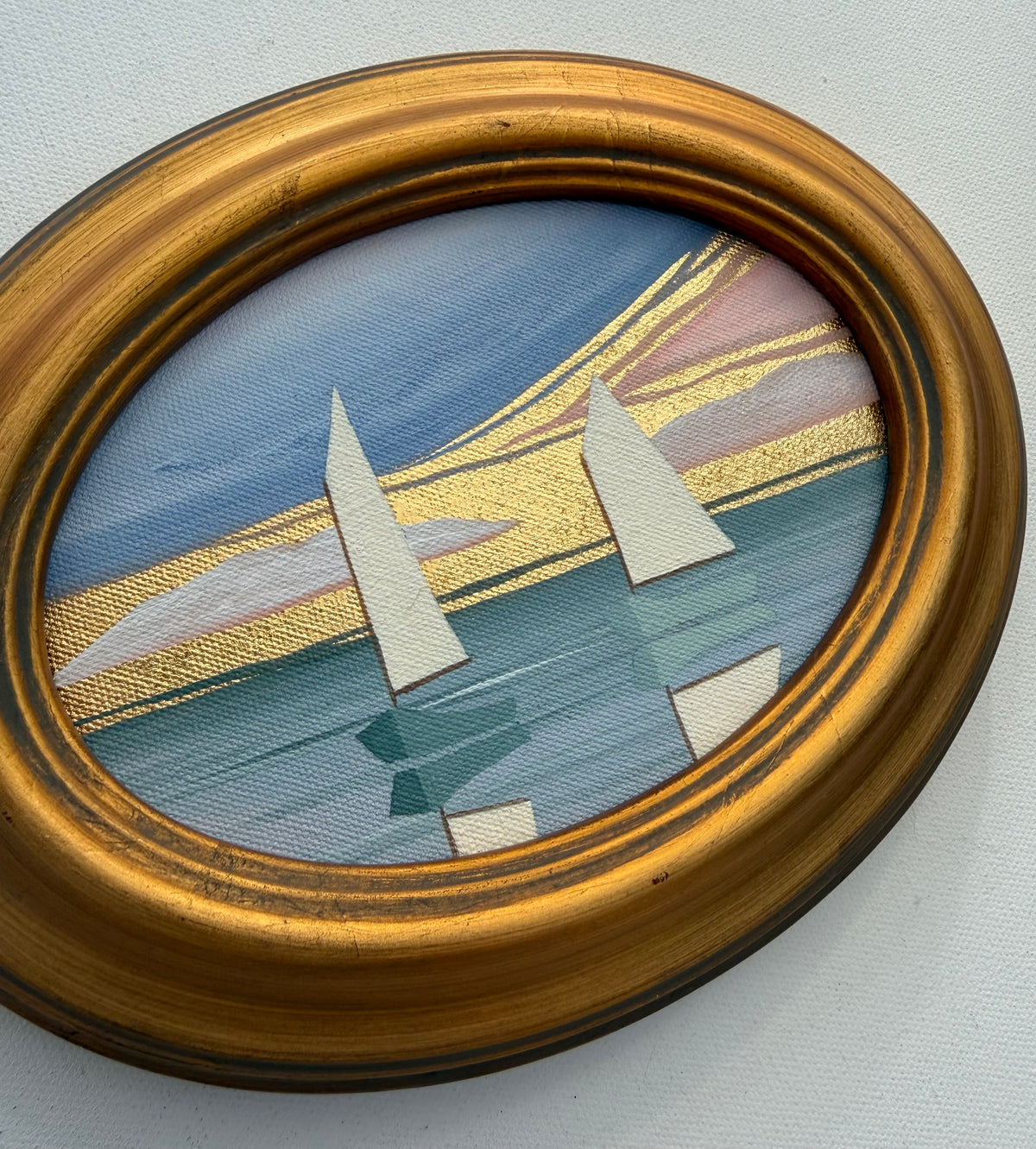 Oval Nikki Storer coastal artwork of sailboats on water with gold leaf accents in a gilded frame.