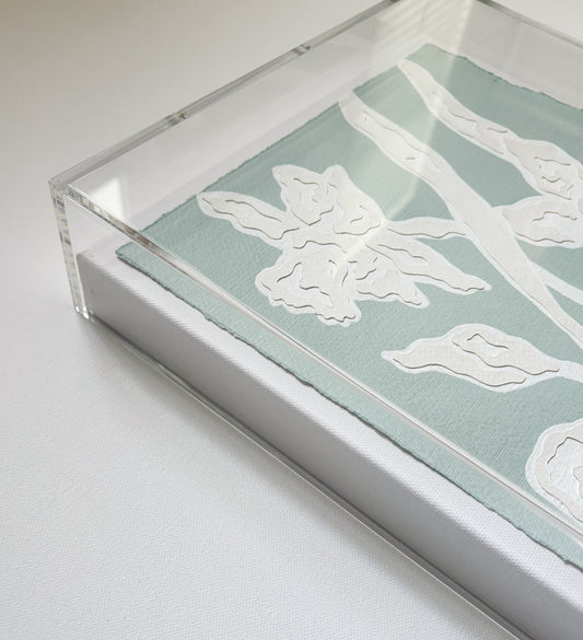 A Kendal Blanchard Art collaged botanical on cotton rag in a clear acrylic frame rests on a white surface.