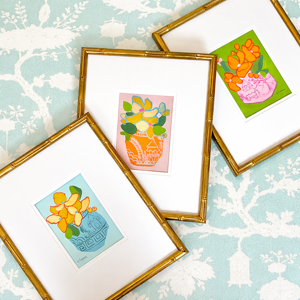 Three colorful floral prints by Kristin Cooney Studio in gold bamboo frames on a light blue patterned surface.