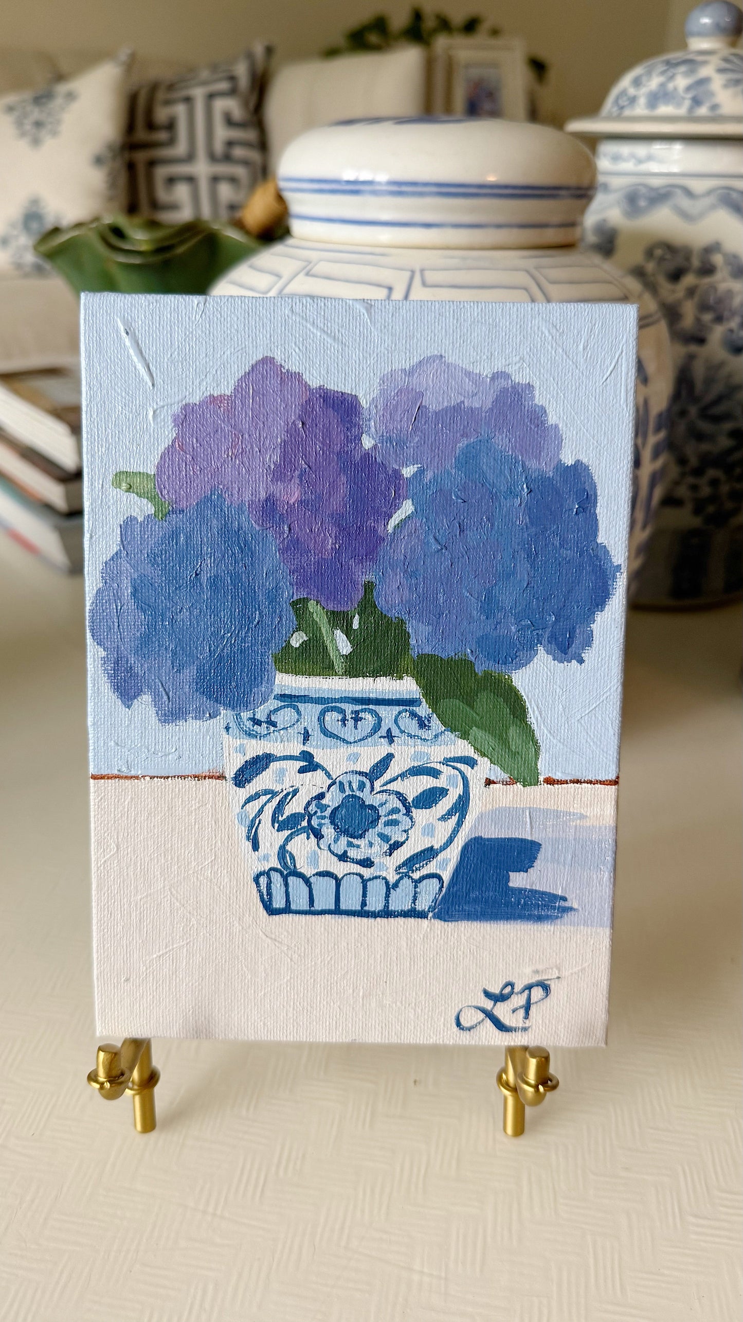 An original Lauri Prins acrylic of purple hydrangeas in a blue-and-white vase with pottery in the background.