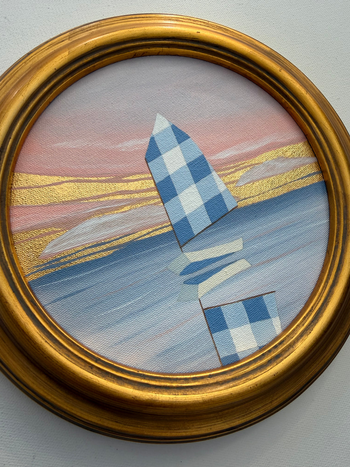 A circular coastal artwork of a blue checkered sailboat on water with 24K gold leaf accents, encased in a gold frame.
