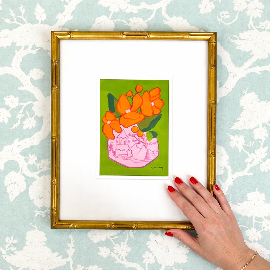 A hand with red nails holds a gold bamboo frame by Kristin Cooney Studio featuring chinoiserie floral art.