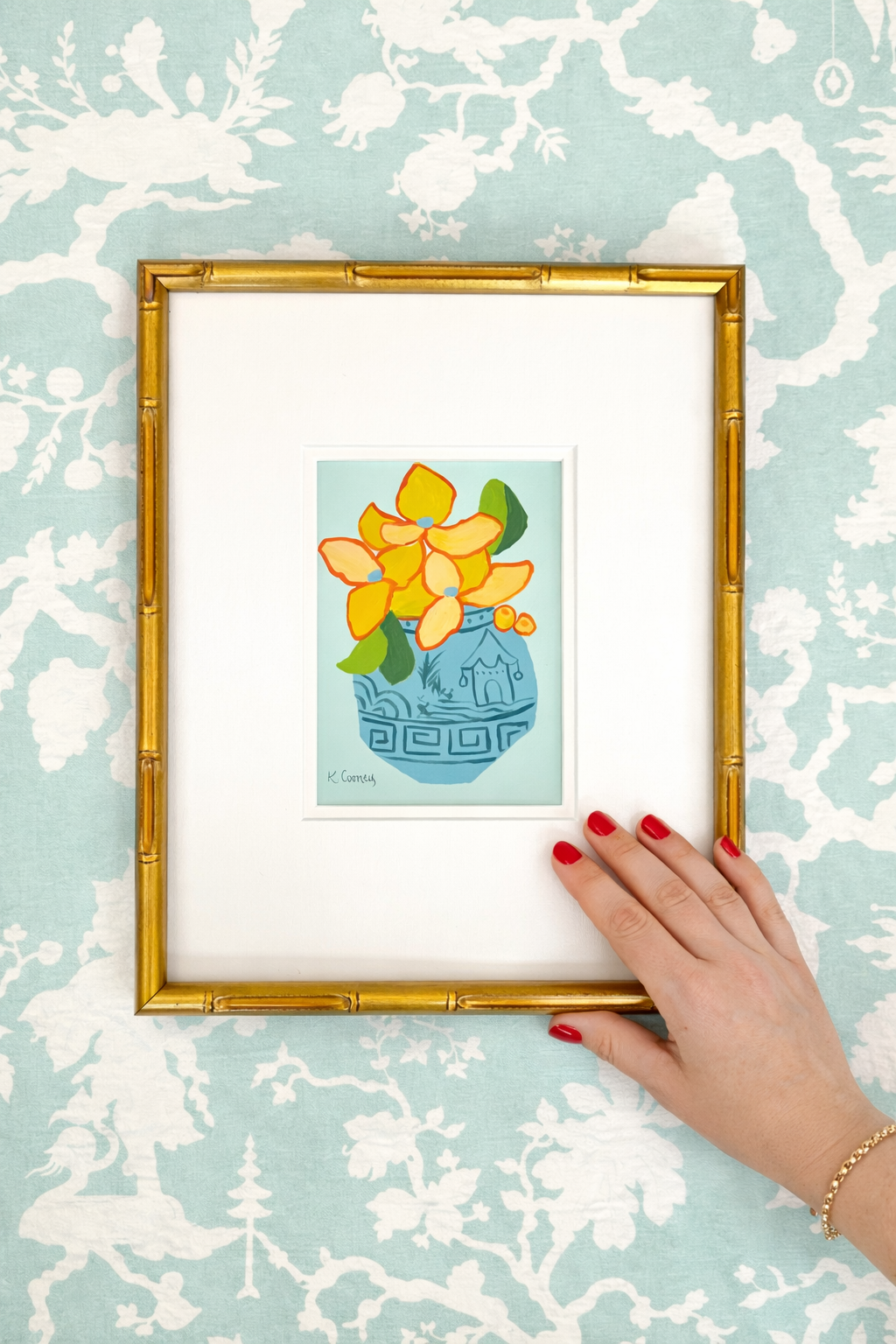A hand with red nails holds a Kristin Cooney Studio gold bamboo frame with chinoiserie yellow flowers artwork.