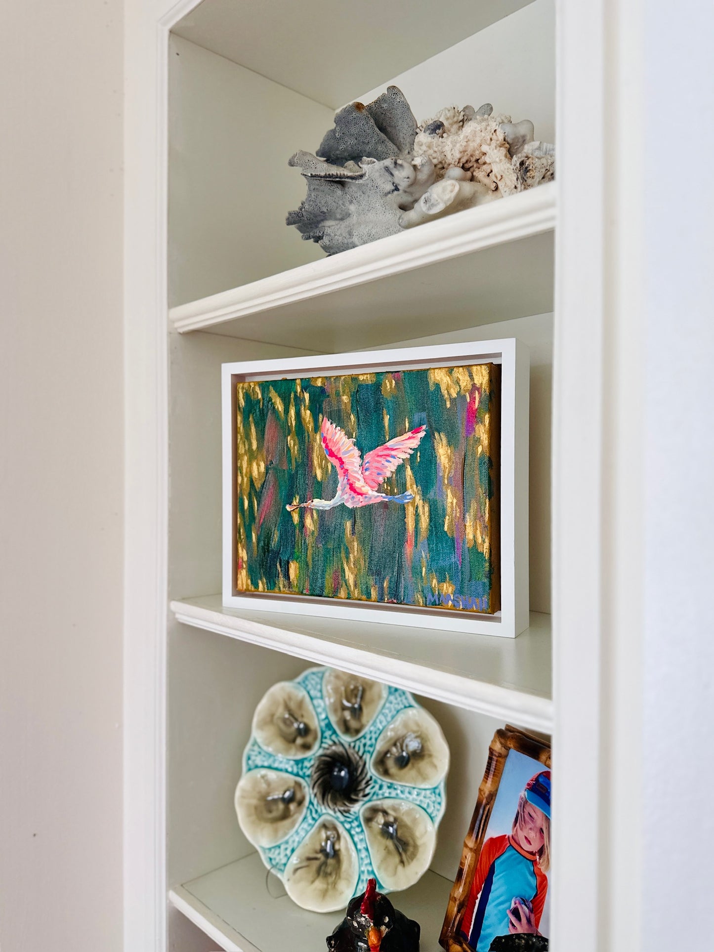 A Lauren Marshall shelf displays coral, a ceramic flower, and a spoonbill painting beside framed Mother’s Day art.