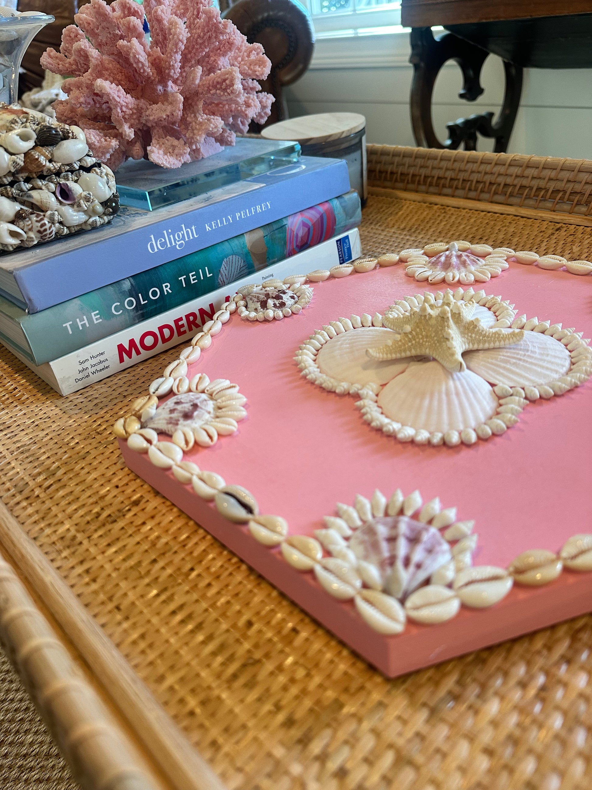 A pink, shell-decorated Kelsey Nichols Art tray sits on a wicker table beside Sailors Valentine books and coral.