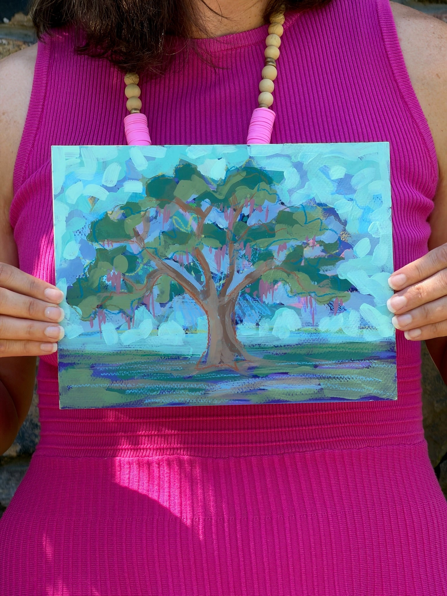 A person in a pink dress holds a watercolor and acrylic live oak tree painting by Elizabeth Sheats Art & Design.
