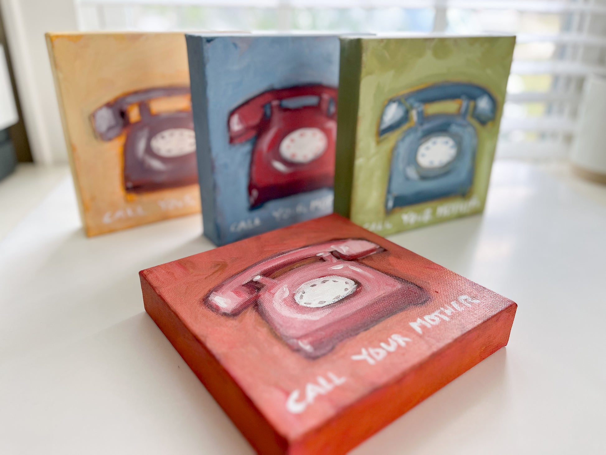 Four Christy Holmes canvases feature hand-painted vintage rotary phones and “Call Your Mother” art on white.