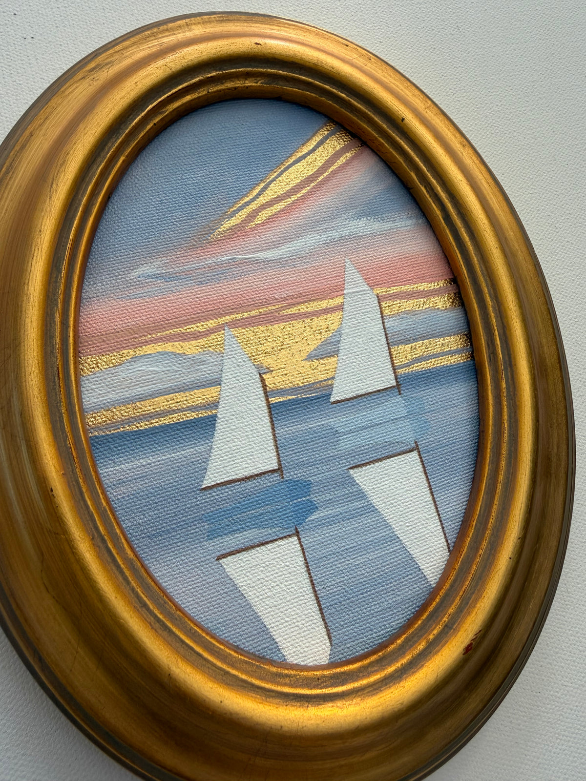 Oval Nikki Storer painting of sailboats on water at sunset with gold accents in a gold frame.