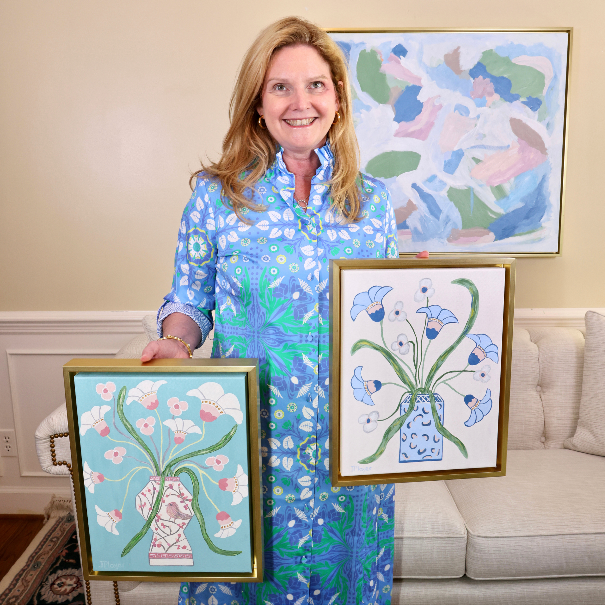 Woman in blue dress smiles, holding two original acrylic paintings in gold float frames with floral art in the living room.