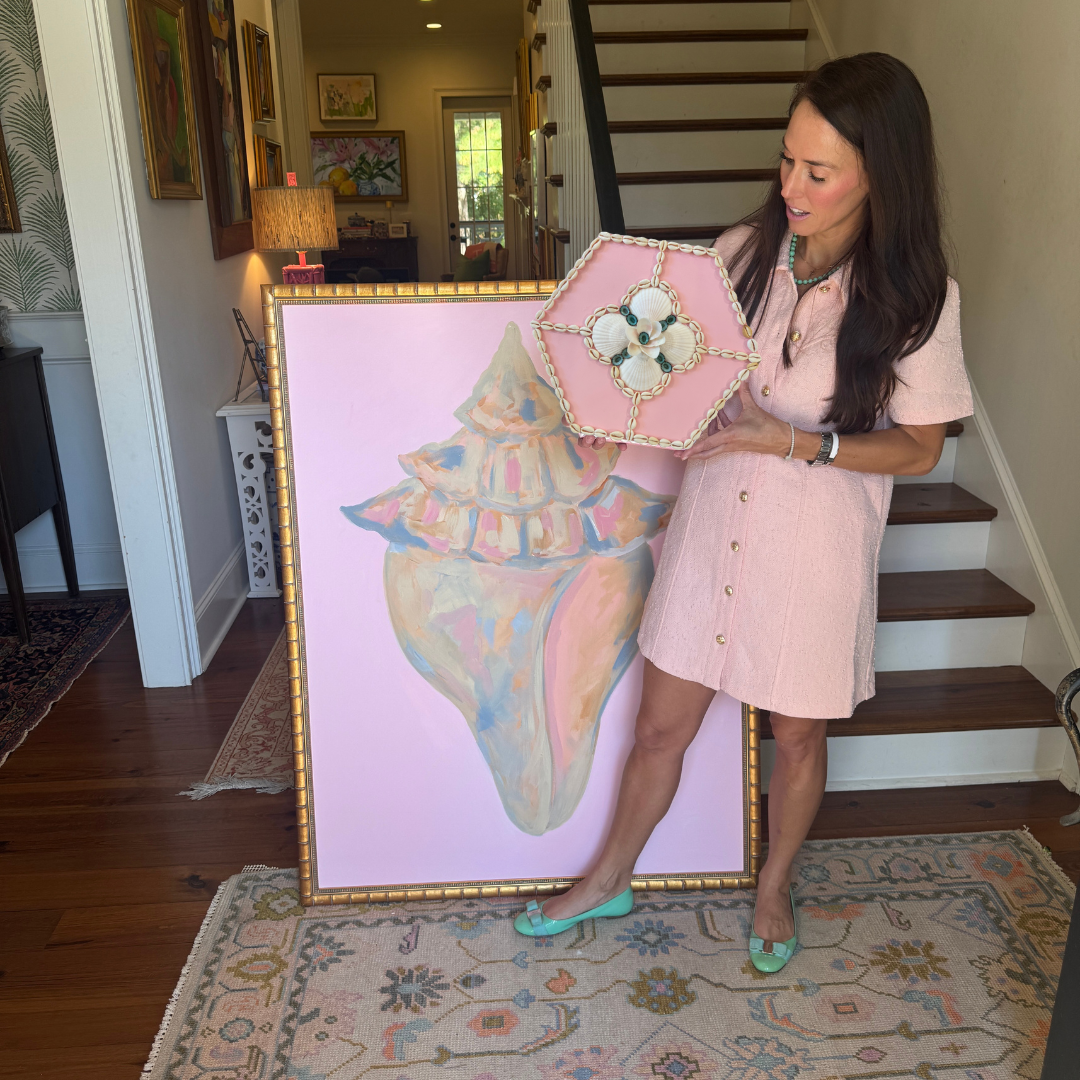 A woman in a pink dress holds shell art by Kelsey Nichols Art, standing beside a Sailors Valentine indoors.