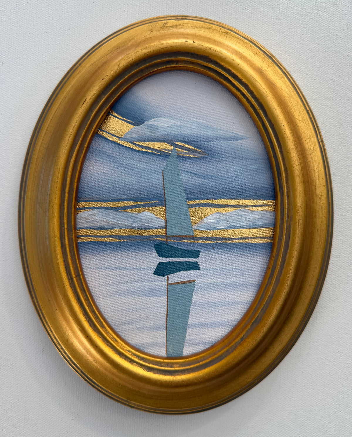 An oval goldleaf painting of a minimalist blue boat on water under gold-painted clouds, housed in a gold frame.