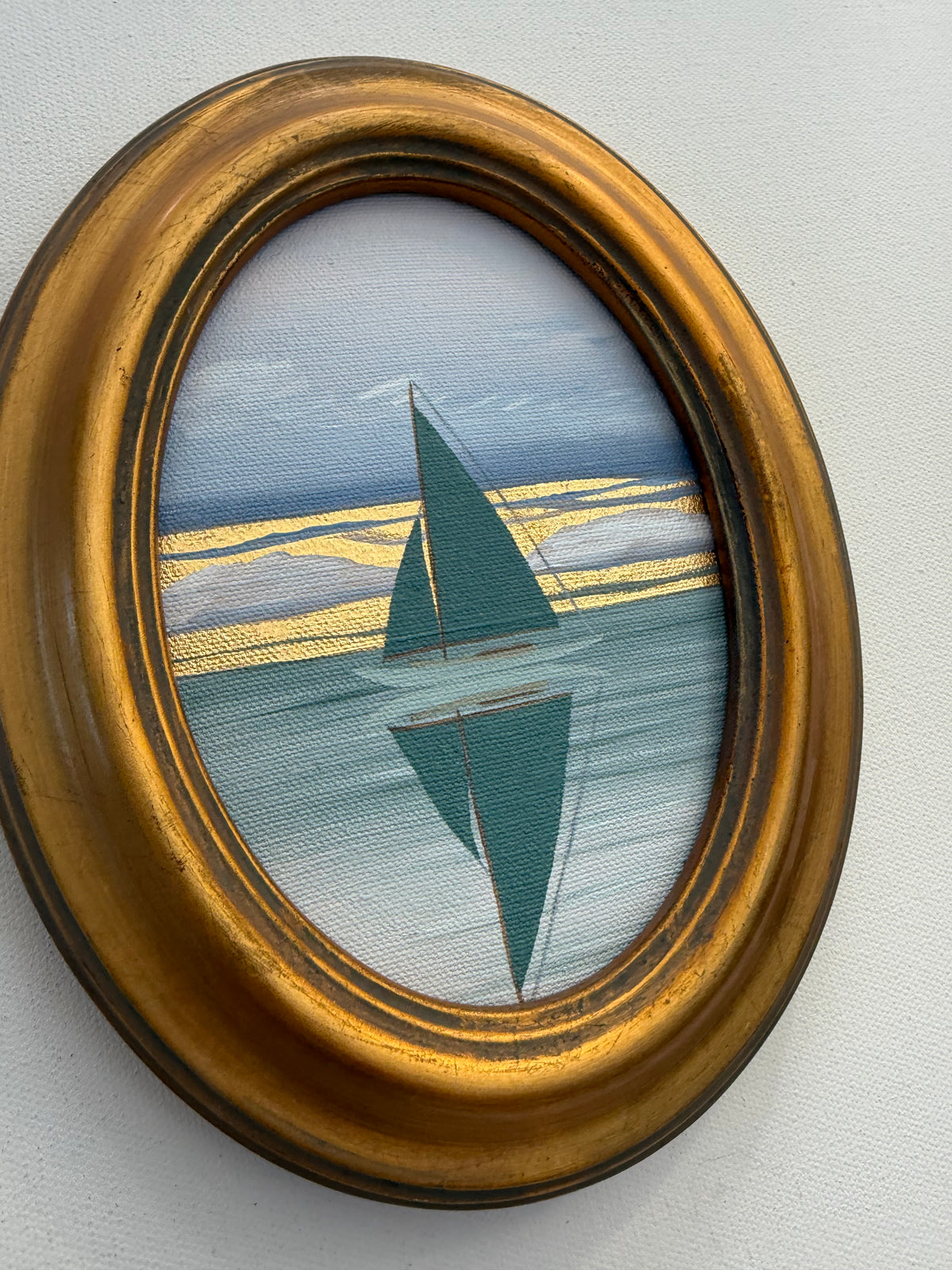 An oval Nikki Storer abstract realism painting shows a teal sailboat reflecting on calm water under a gold-streaked sky.