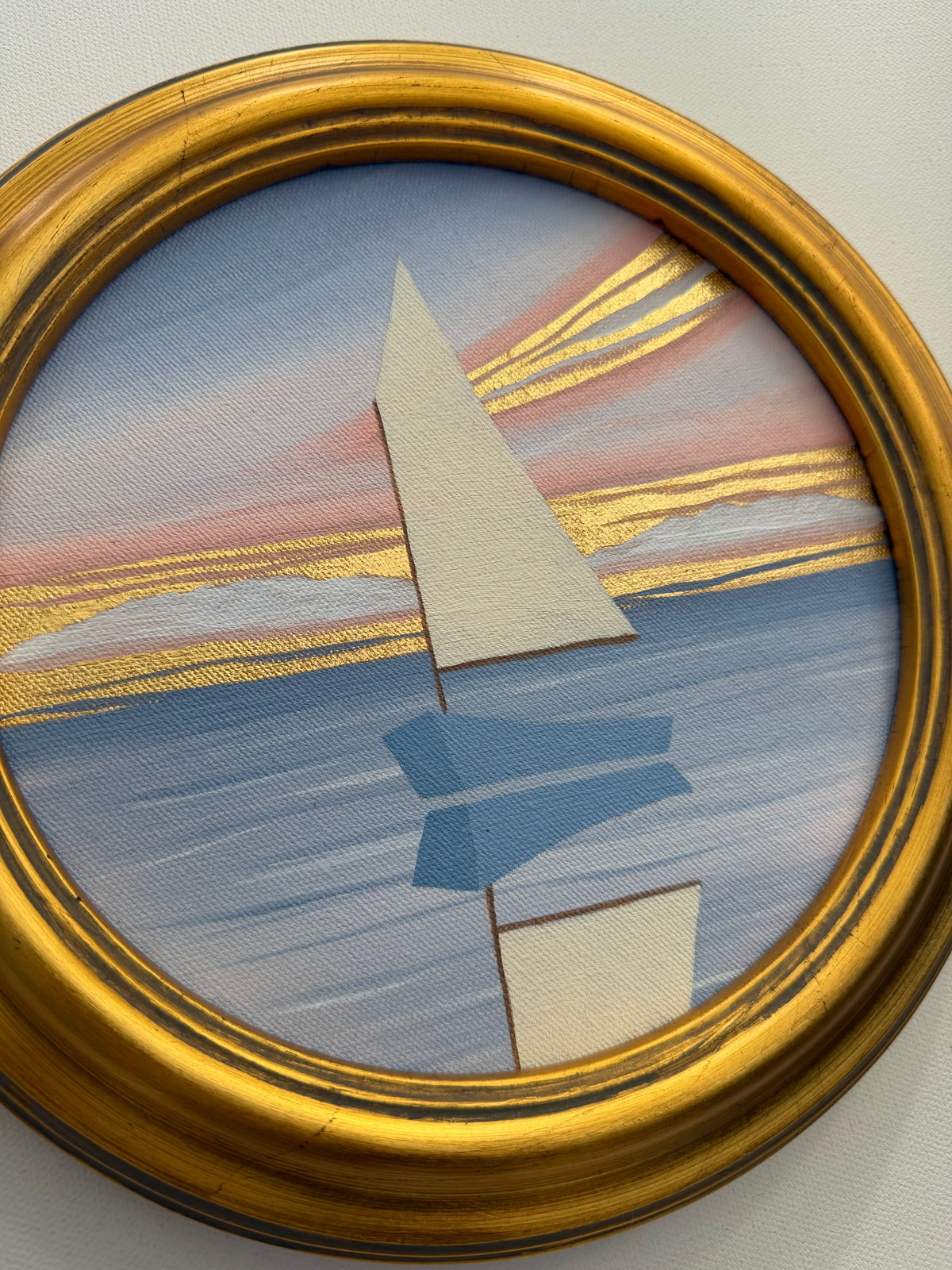 A circular coastal painting of a stylized sailboat on water at sunset with 24K gold leaf accents, in a gold frame.