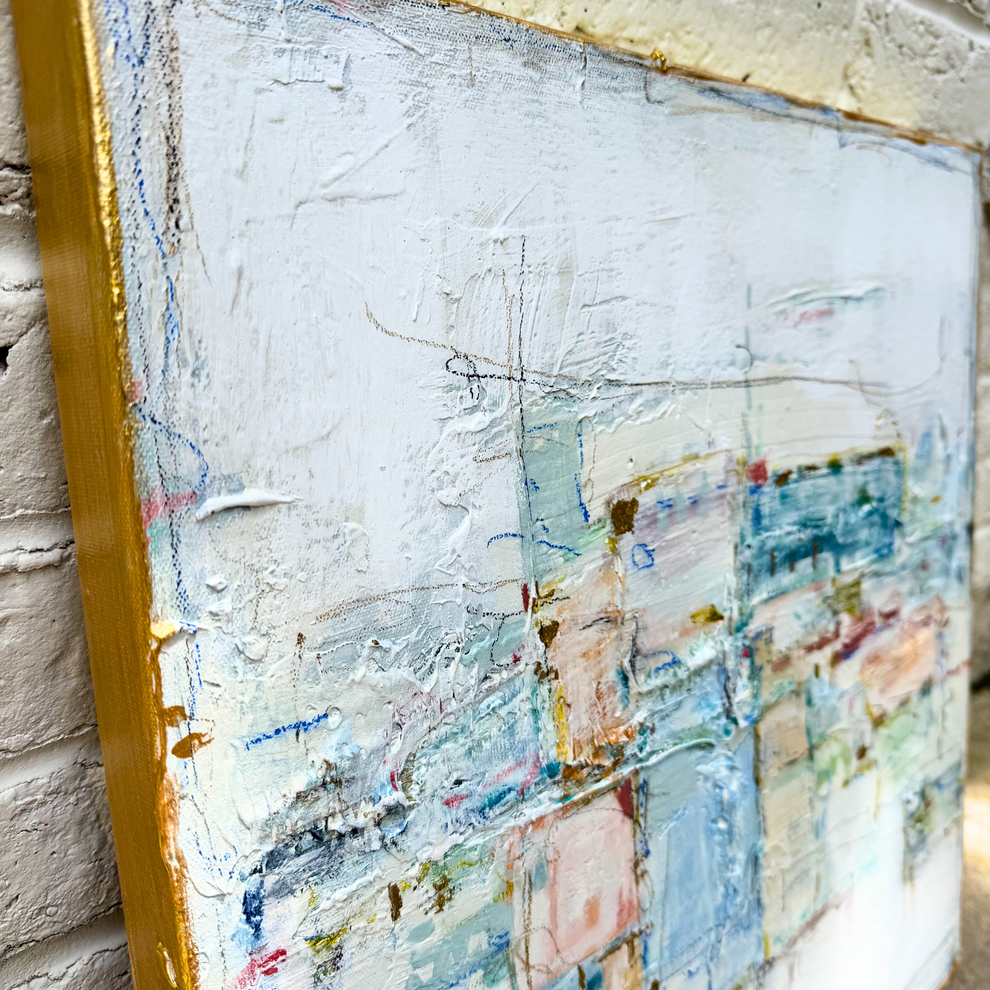 Close-up of a Megan Guidry abstract painting in blue pastels, set in a gold frame against a brick wall.