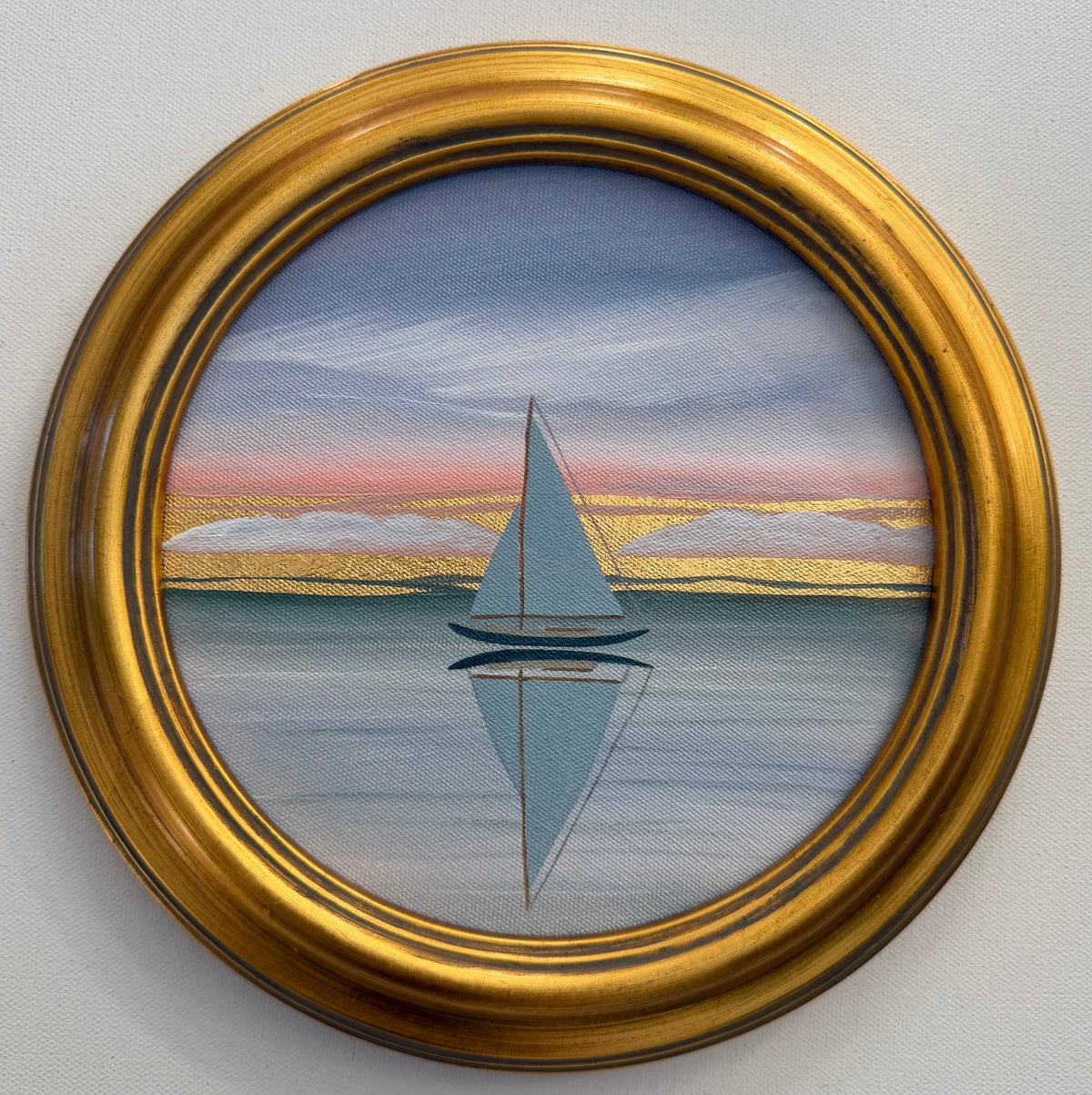 A circular gold-framed coastal artwork of a sailboat on calm water reflecting a sunset sky with 24K goldleaf clouds.