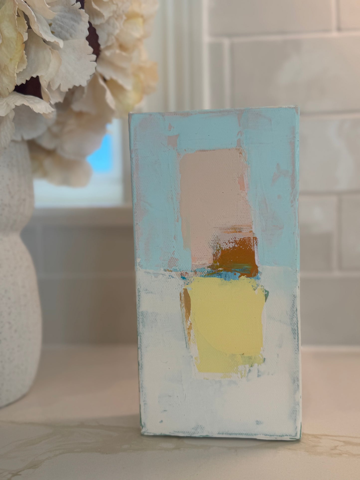 KP Art Collective abstract painting features pastel Mediterranean blocks on a light background beside a floral vase.