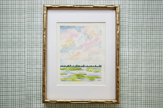 Framed watercolor painting of a landscape with colorful clouds on a checkered wall.
